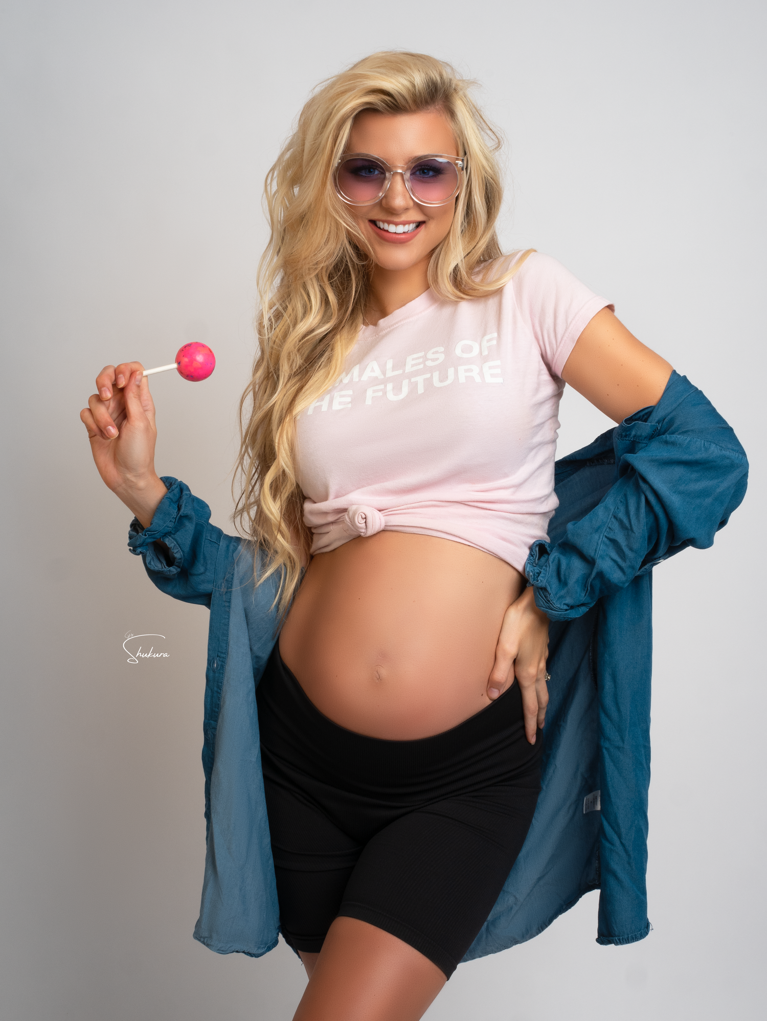 A pregnant woman in a pink t-shirt and blue denim jacket holds a pink lollipop while posing against a gray background.