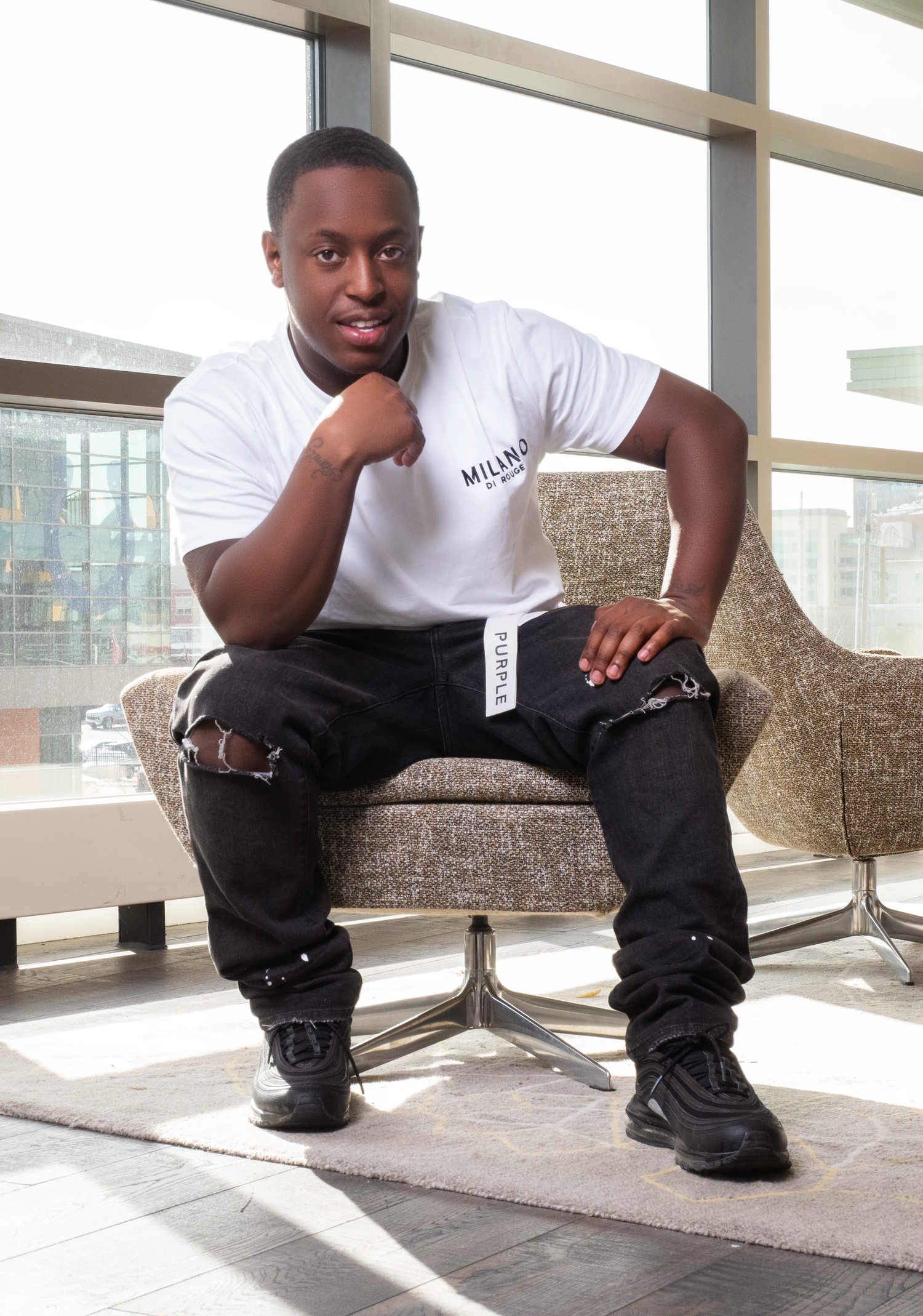 A person in a white t-shirt and black pants sits relaxed on a grey upholstered chair near large windows in a bright space.