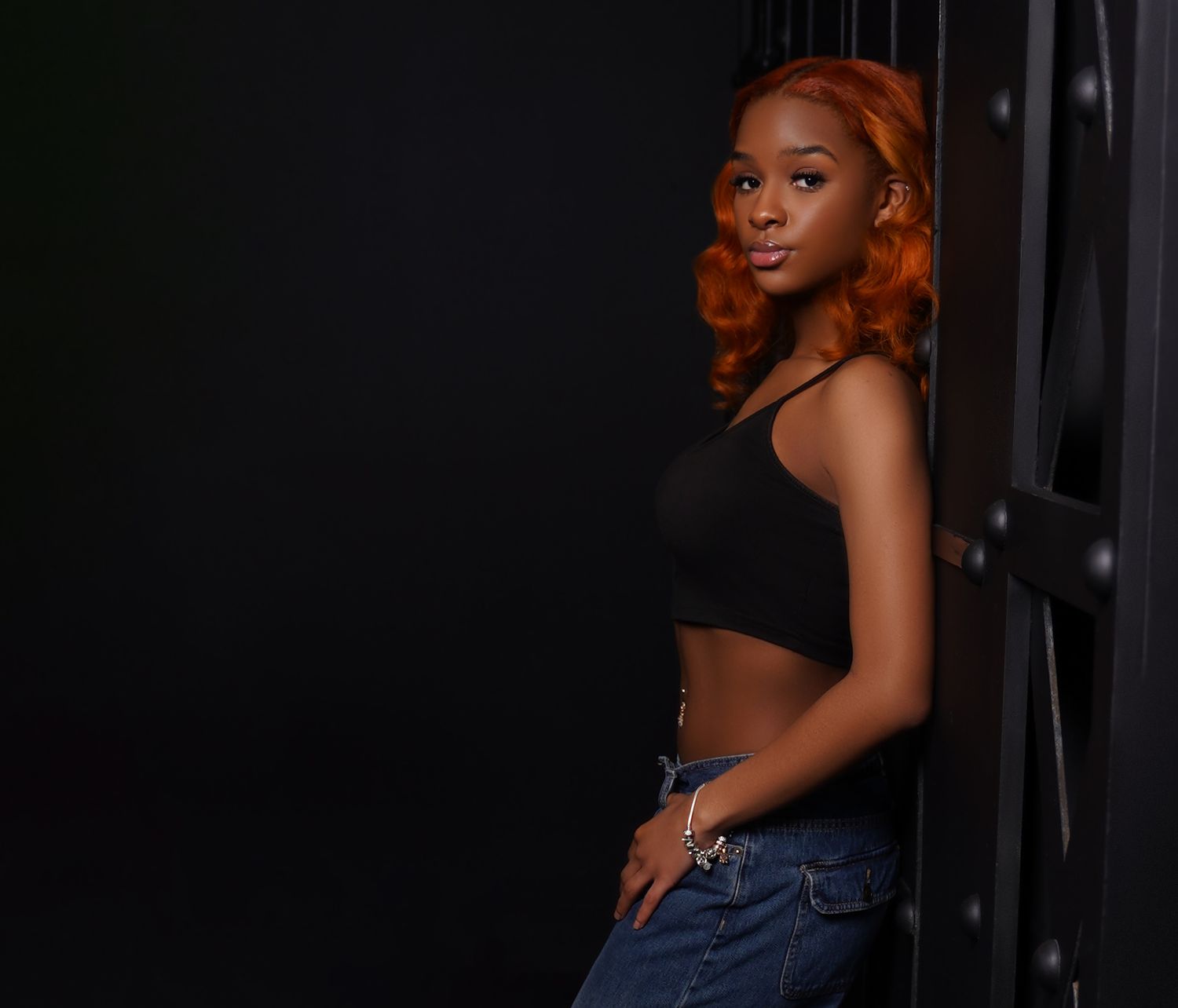 Someone with vibrant copper-red curls poses confidently in a black crop top and jeans against a dark background.