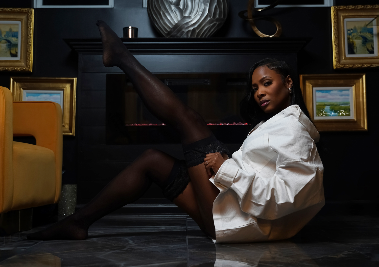 A dancer in a white dress and socks poses elegantly on wooden steps with framed artwork in the background.