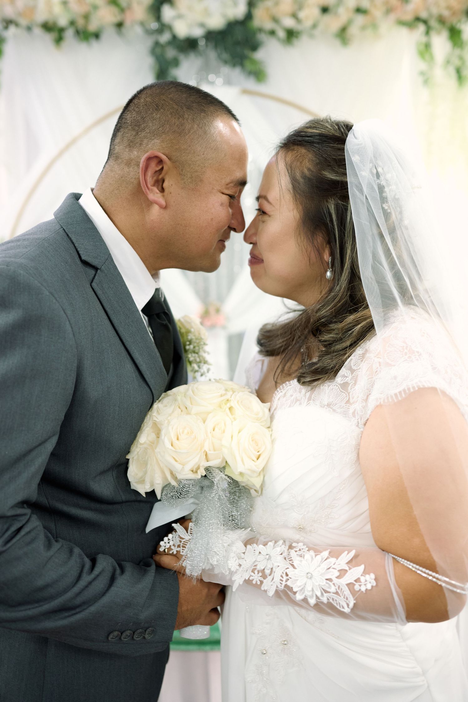 A tender moment between newlyweds sharing a romantic kiss under floral decorations, with the bride holding white roses.
