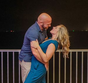 A romantic couple embraces on a balcony overlooking city lights at night, with the woman in a blue dress.