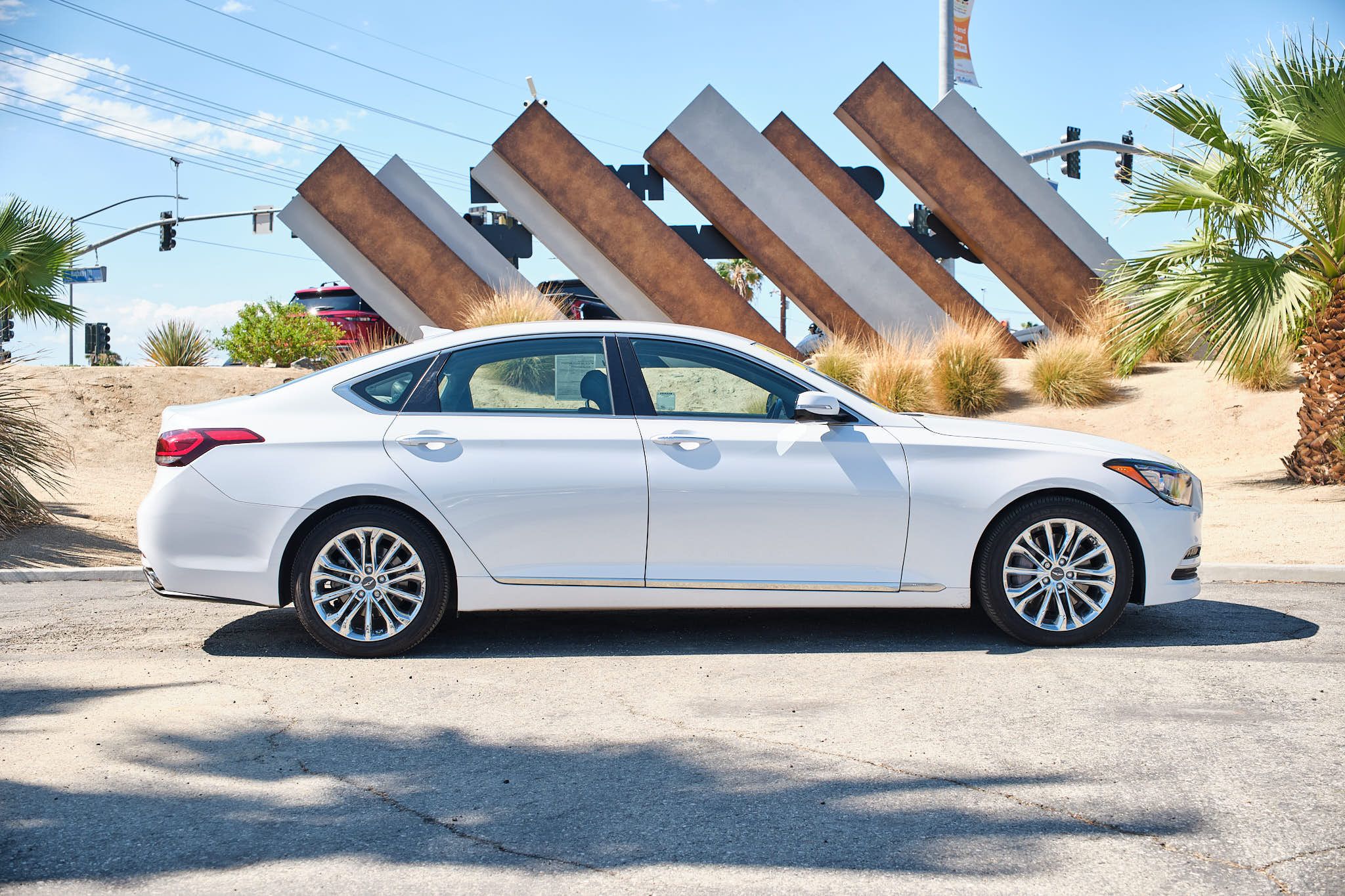 White luxury sedan parked in front of angular metal art installation with palm tree in background.