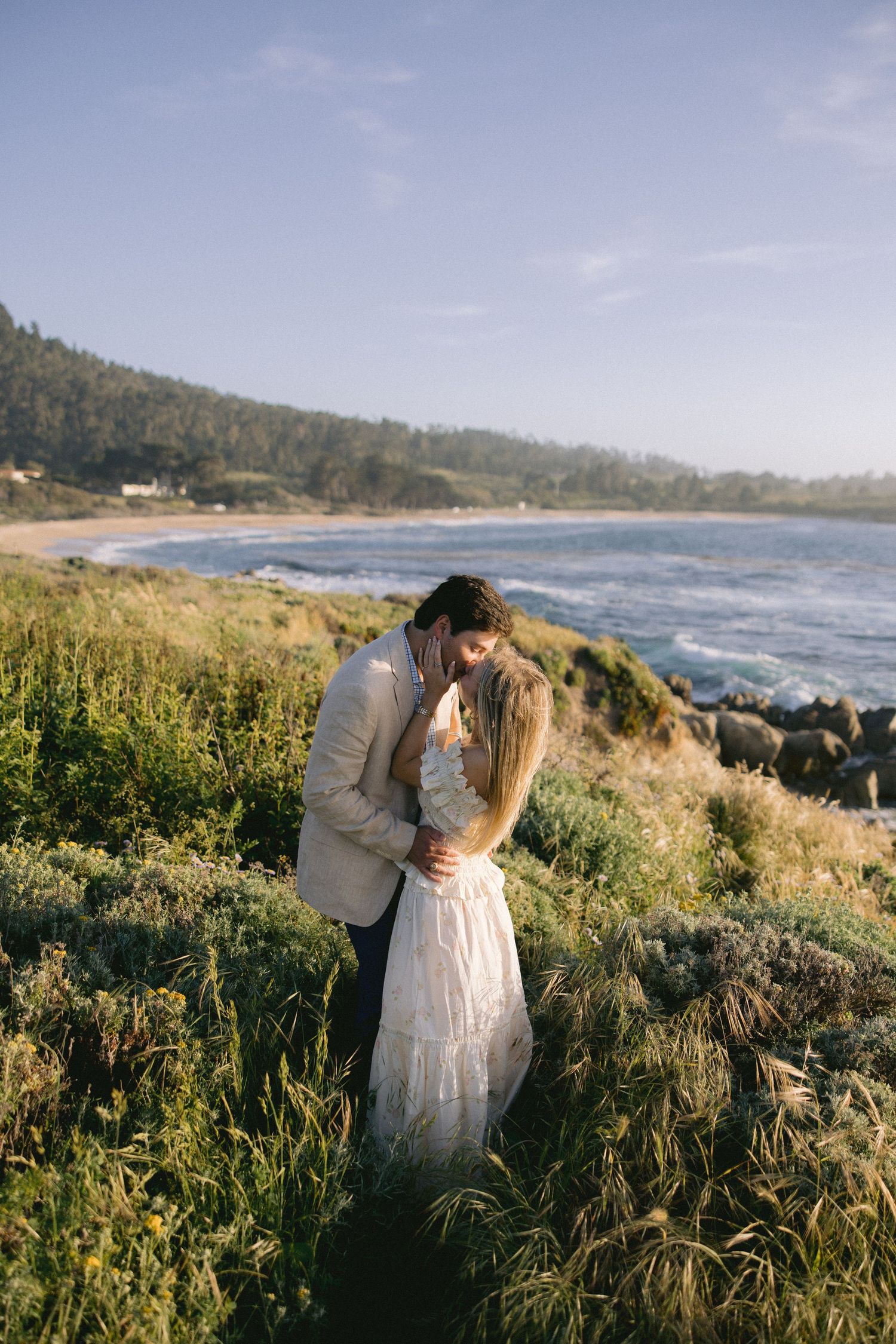 A couple embraces on a scenic coastal cliffside with tall grass and ocean waves in the background during golden hour.