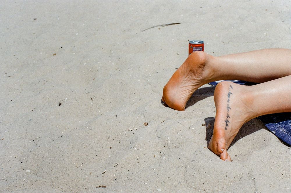 Feet with small ankle tattoos resting on sandy beach against bright sunlight.