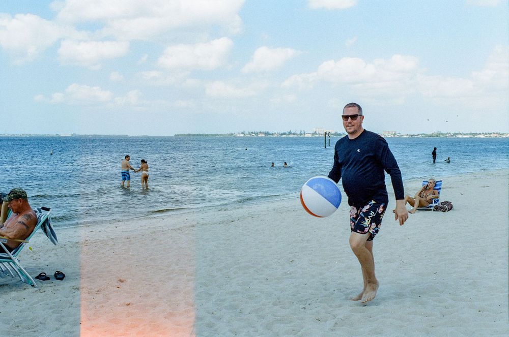 A sunny day at a beach with people enjoying the sand and water while a beachgoer carries a white volleyball.