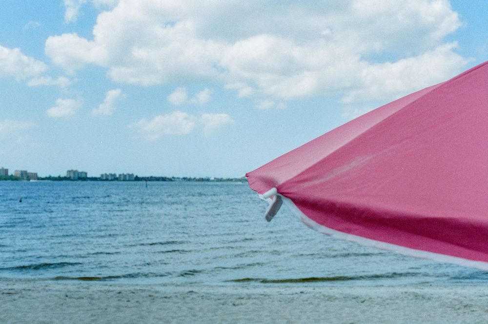 Pink beach umbrella overlooking a serene blue lake on a partly cloudy summer day.