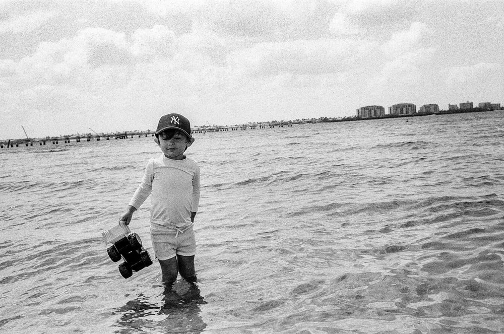 A small figure stands in shallow ocean waves holding a bucket on a cloudy beach day.