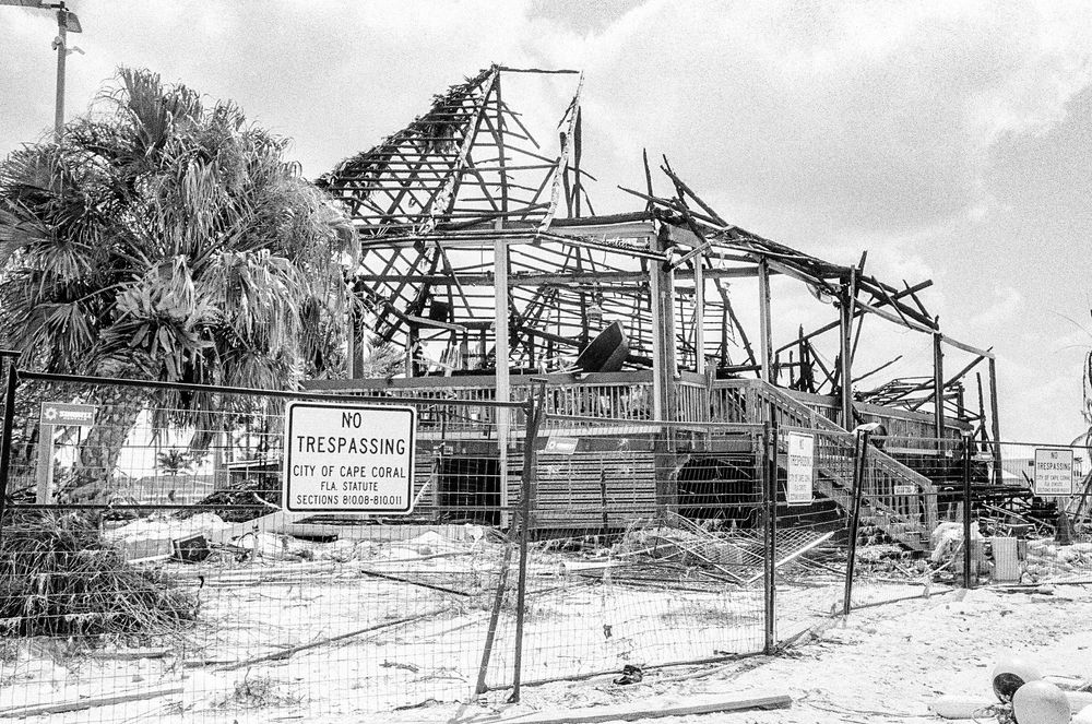 Black and white photograph of a damaged building structure with exposed metal framework behind a chain link fence.