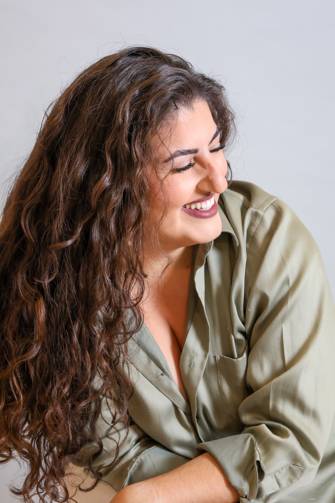 A joyful expression with long dark curly hair wearing a sage green button up shirt against a light background.