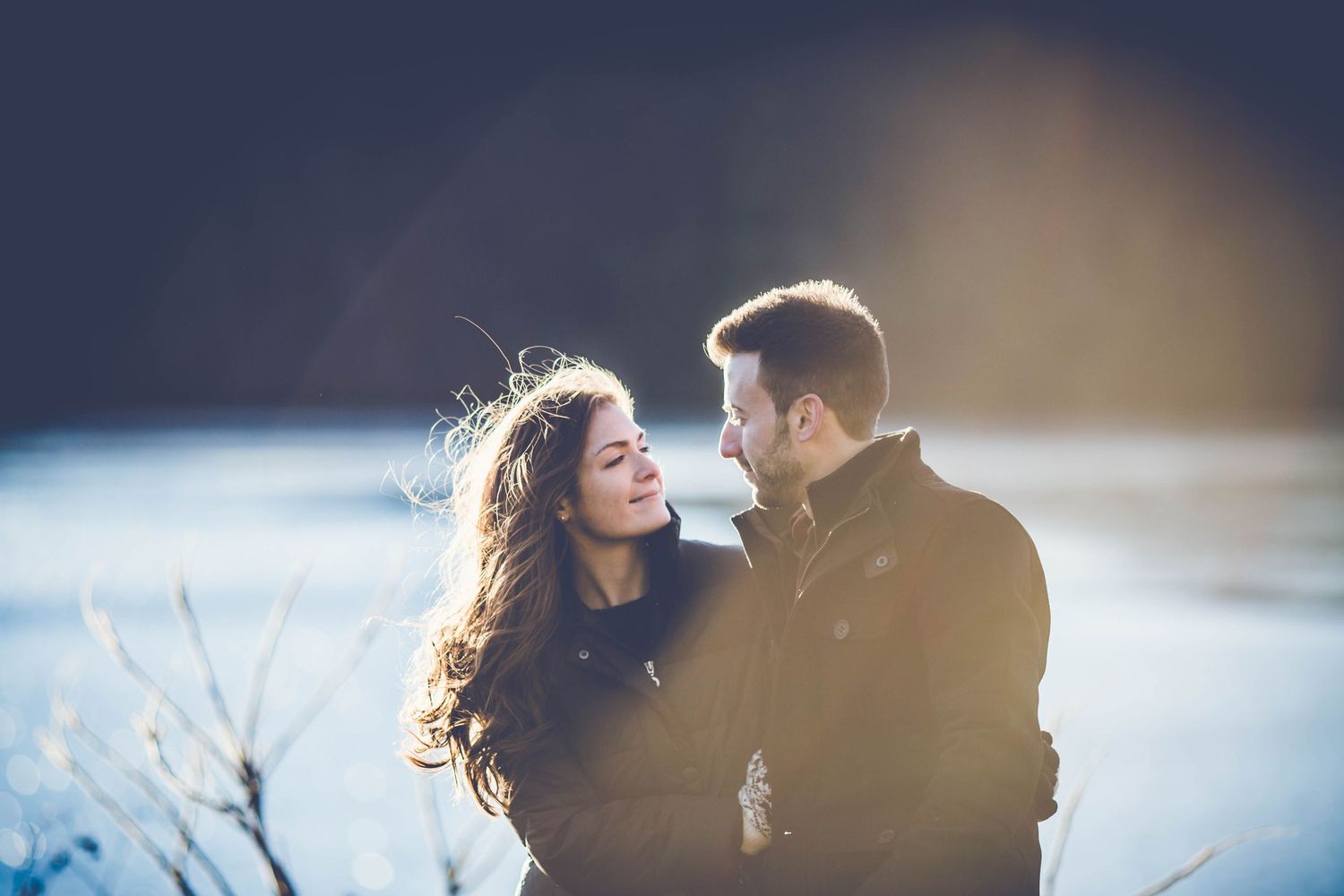 Romantic winter portrait of couple gazing at each other with backlit snow scenery.