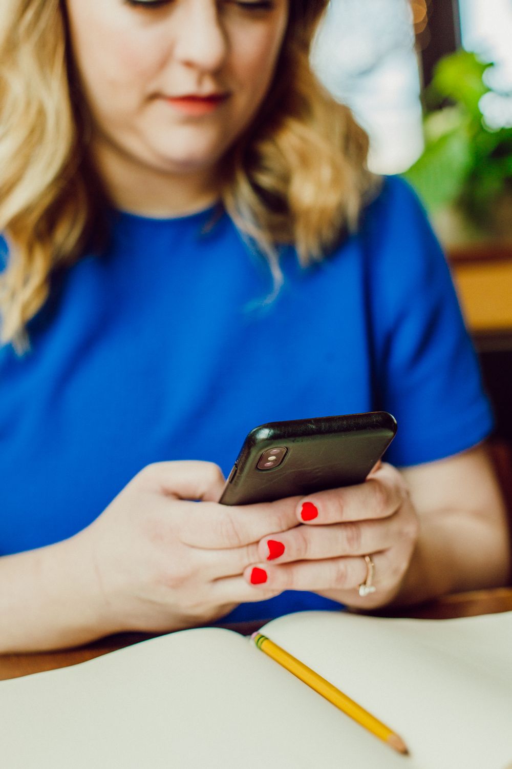 Close-up of hands with red nail polish holding a smartphone over a notebook with pencil.