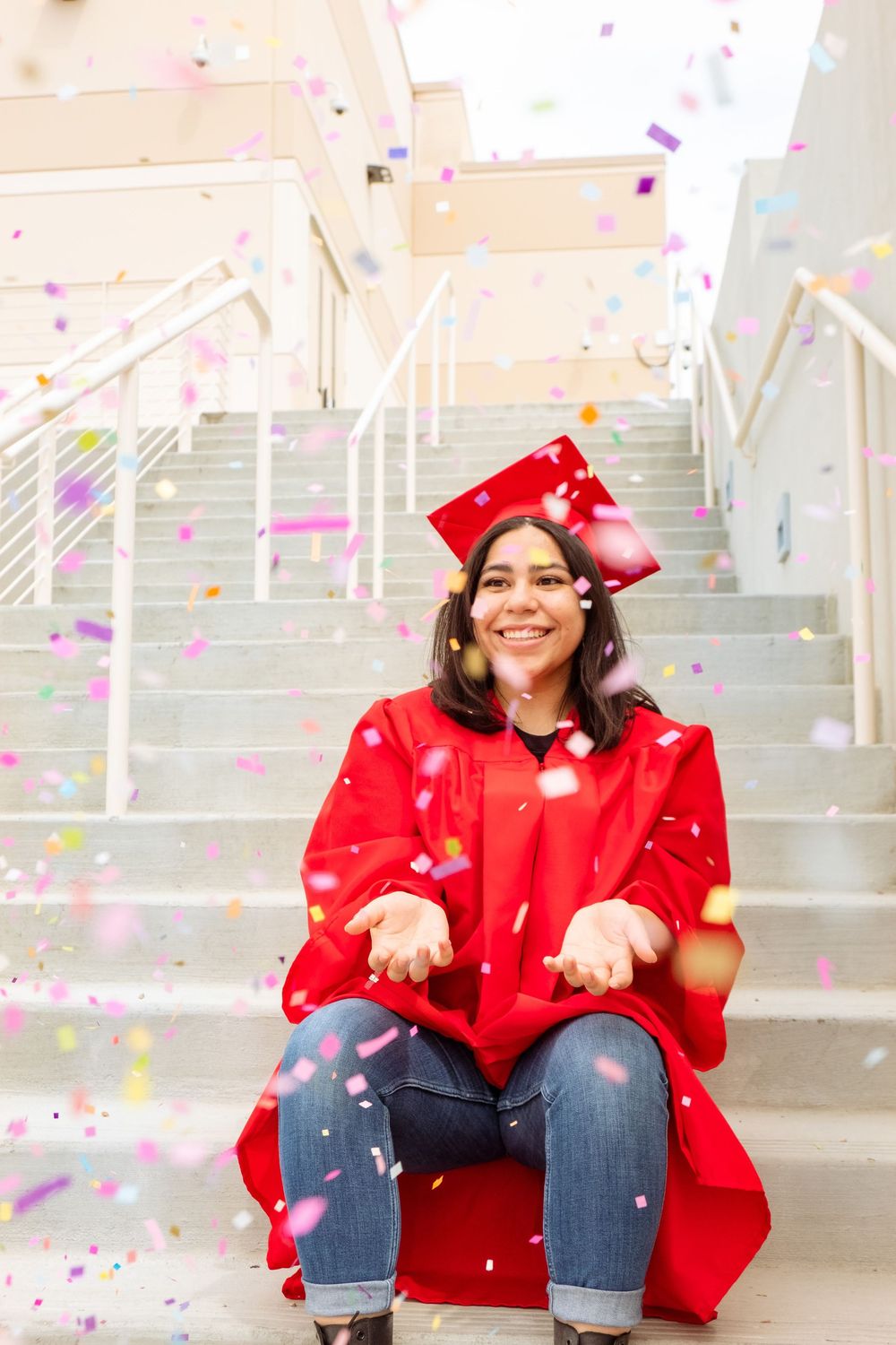 Graduate in red graduation gown sitting on stairs surrounded by falling confetti.
