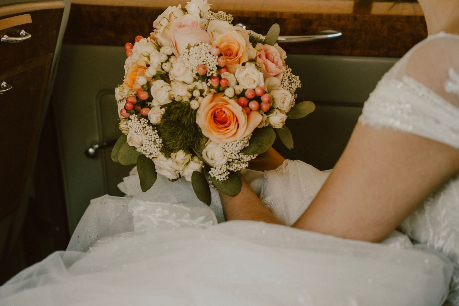 A bridal bouquet with peach roses and white flowers rests on a white dress in a car.