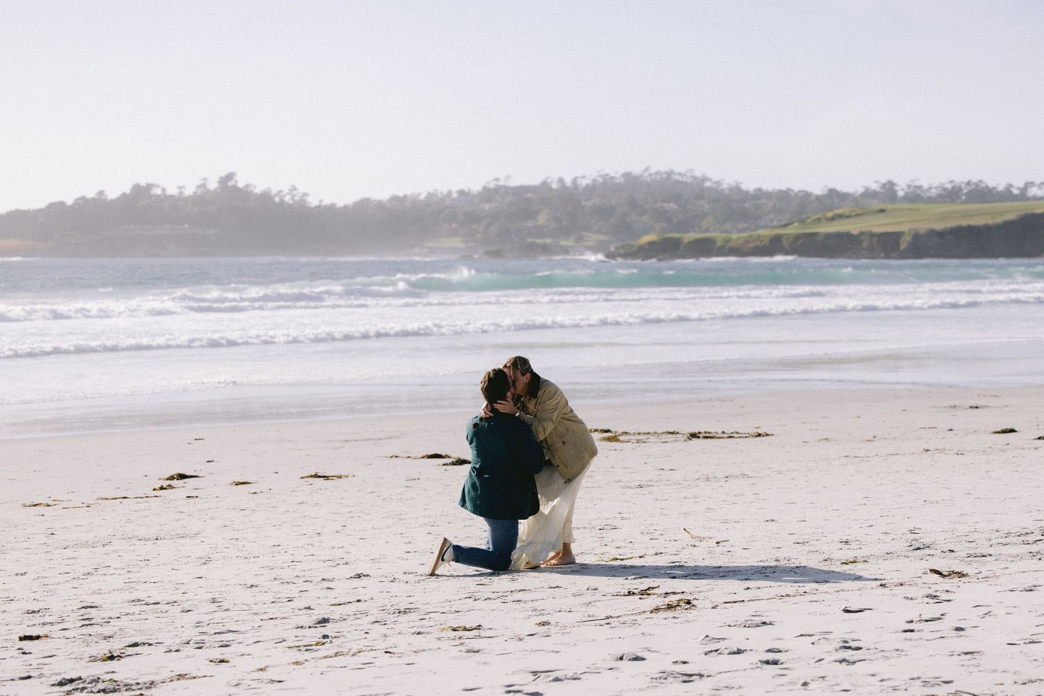 Two people in warm coats share an embrace on a windswept beach with rolling waves and hills in the background.
