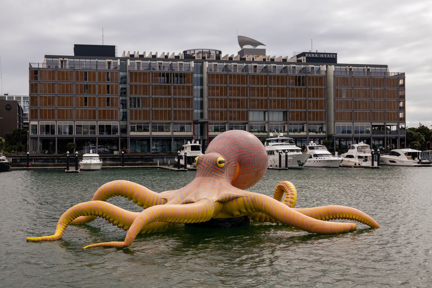 Large inflatable octopus sculpture floating in a harbour, with boats and a modern waterfront building in the background.