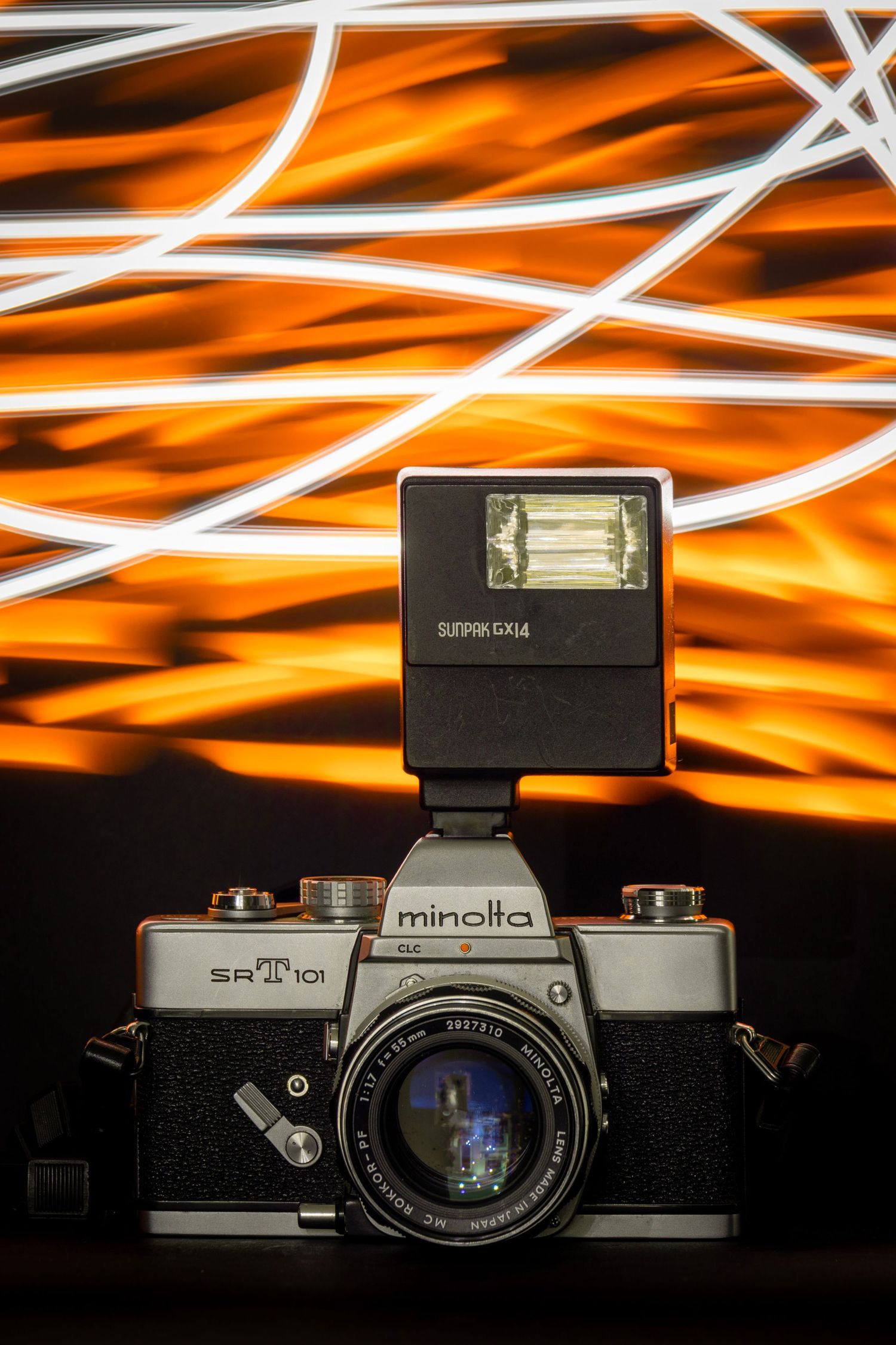 Vintage Minolta film camera with flash attached, set against a background of dynamic white and orange light trails.