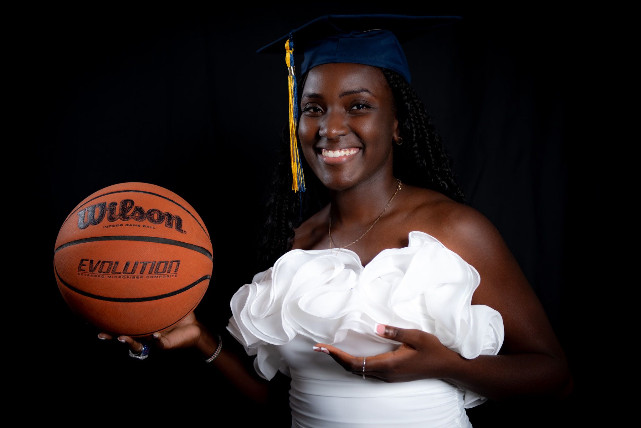 Graduate in white dress holding a basketball and wearing blue cap with gold tassel in a dark studio setting.