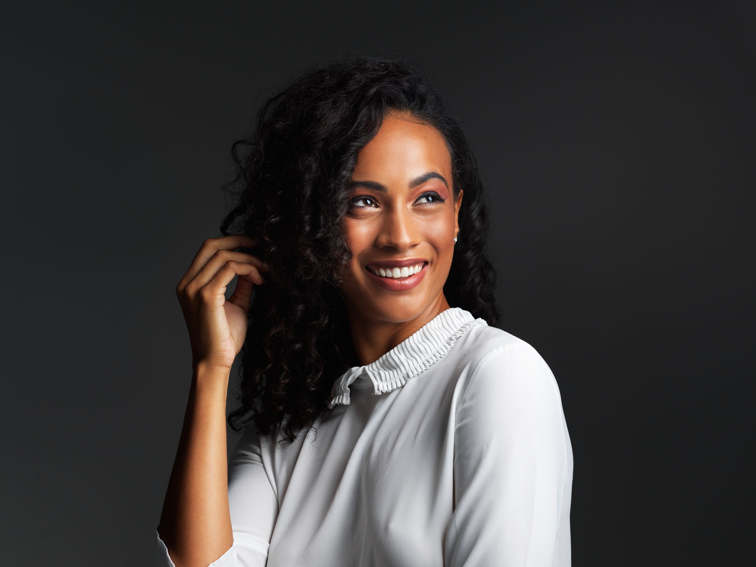 Professional portrait against dark background showing bright smile and white collared shirt while touching curly dark hair.