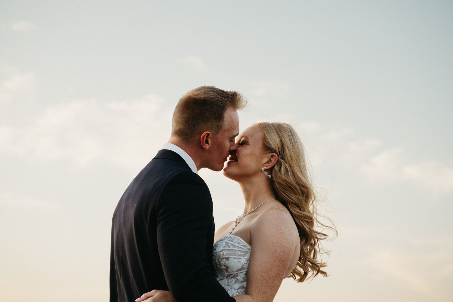 A romantic couple kisses outdoors at sunset, with soft clouds in the background and the bride's curled hair flowing.