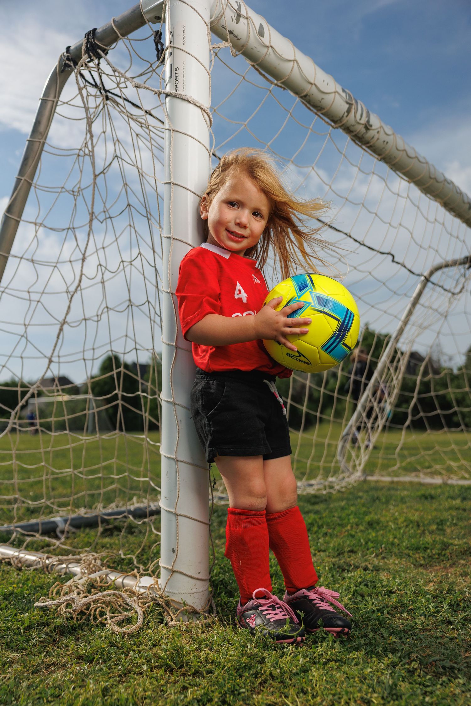Young soccer player in red uniform holds yellow ball while standing by goal post.