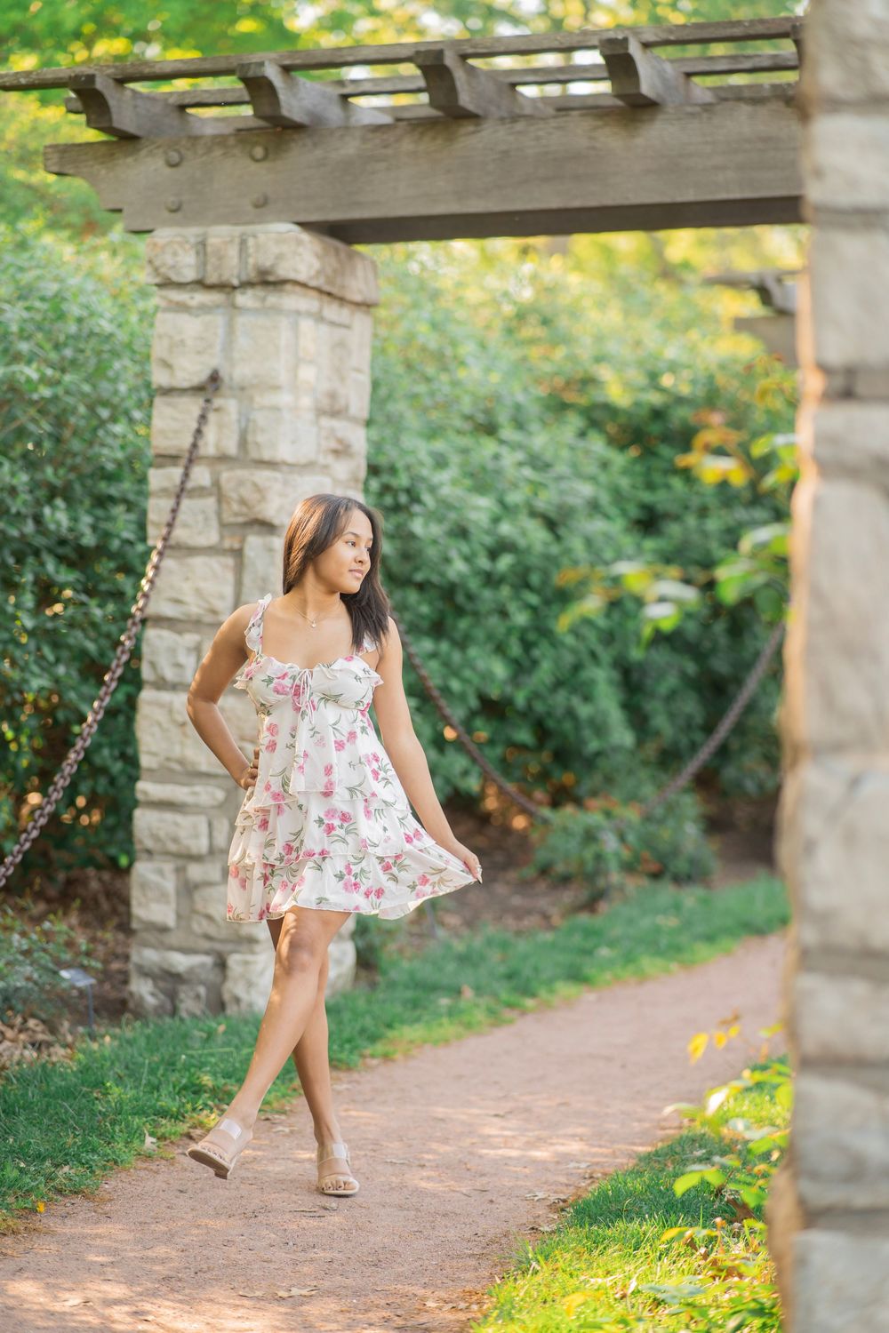 Young woman in floral sundress walking gracefully under stone pergola in garden.