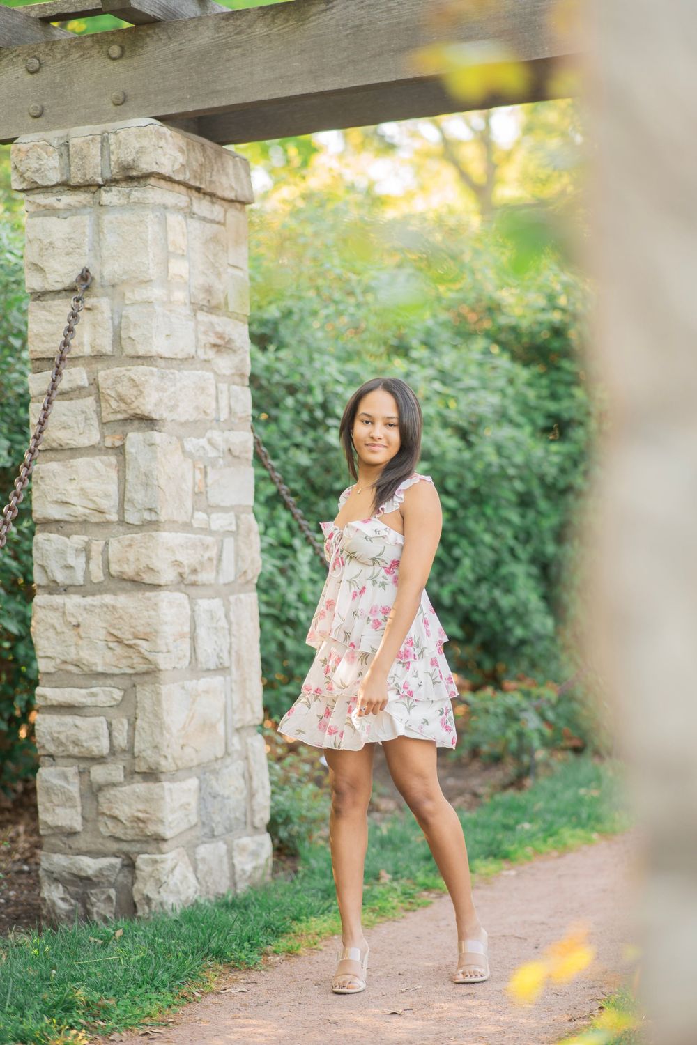 Teen girl in floral dress posing beside stone column on sunlit garden path.