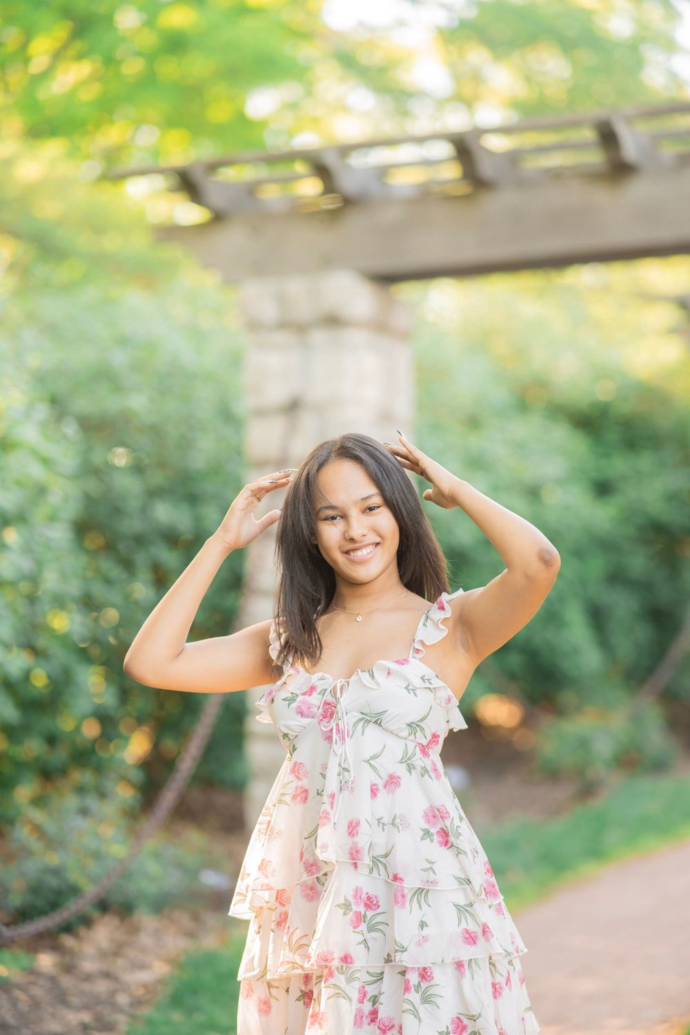 Smiling teen girl in floral dress with hands in hair, standing in garden.