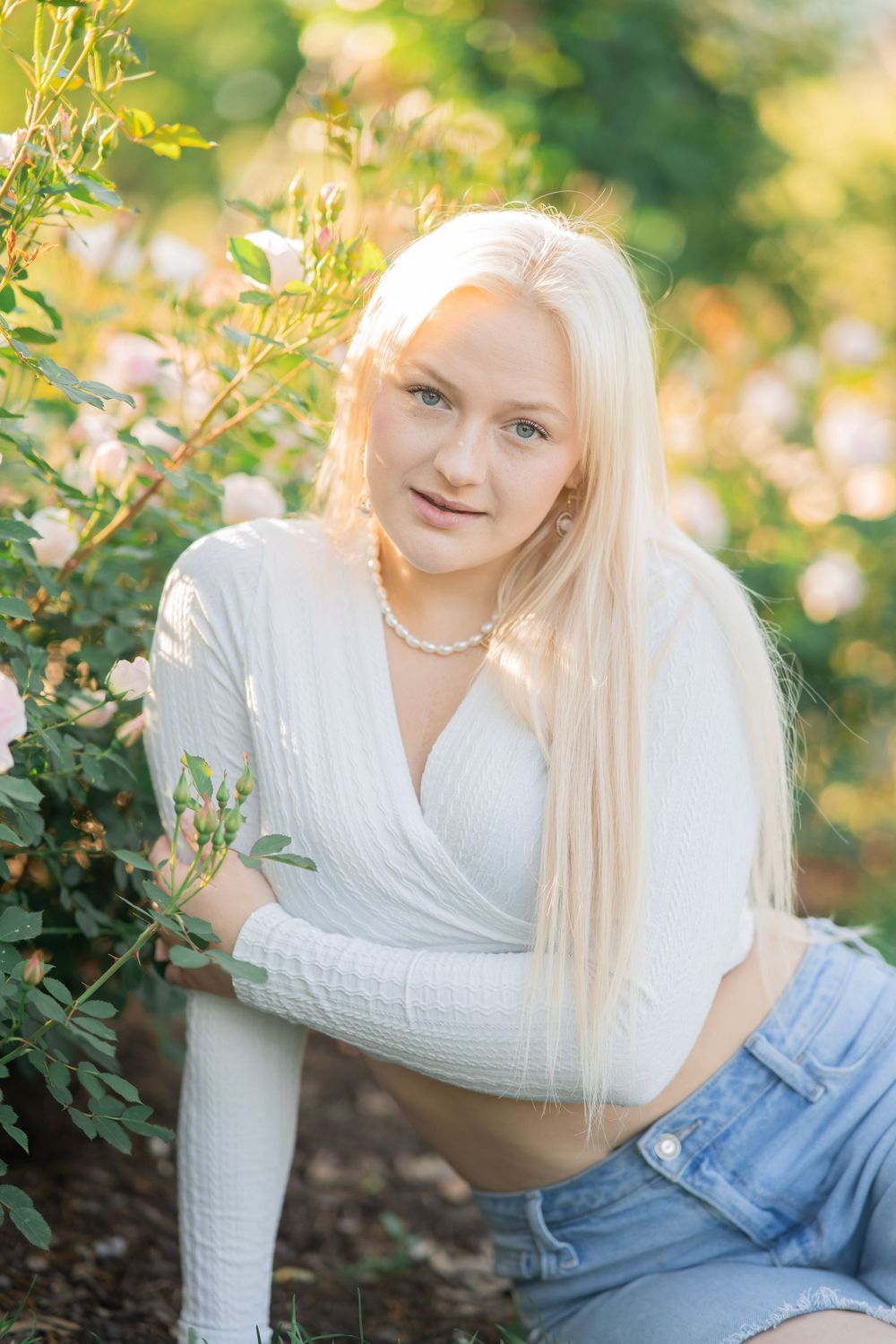 Blonde teen girl in white wrap top and jeans posing near blooming rose bushes.