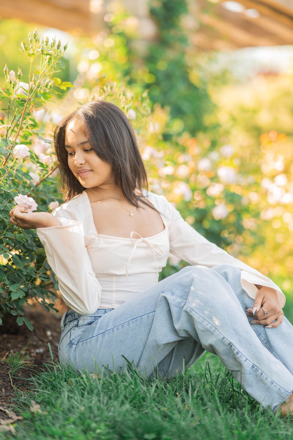 Young woman in white top and jeans sitting on grass admiring white roses in a sunlit garden.