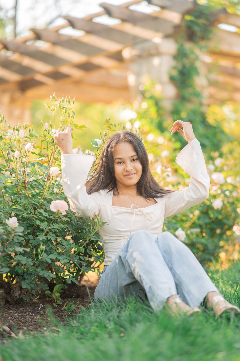 Young woman in white top and jeans sitting on grass flexing arms beside white roses under a pergola.