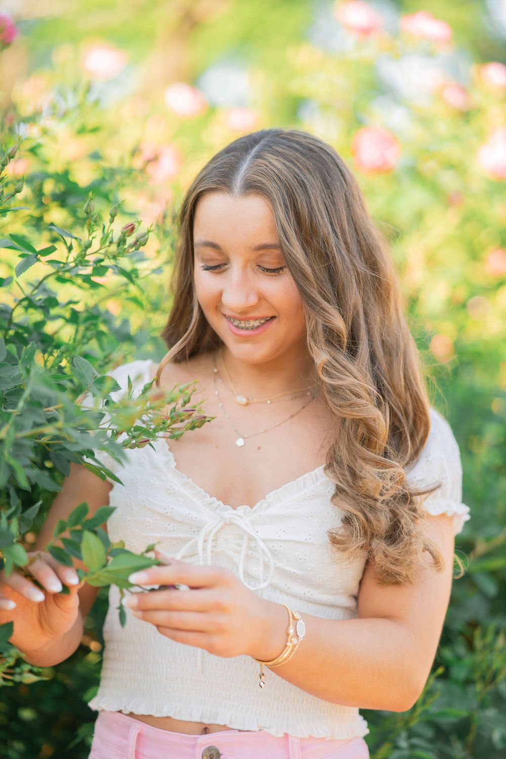 Smiling teenage girl with curly hair in white crop top and pink pants touching green leaves in garden.