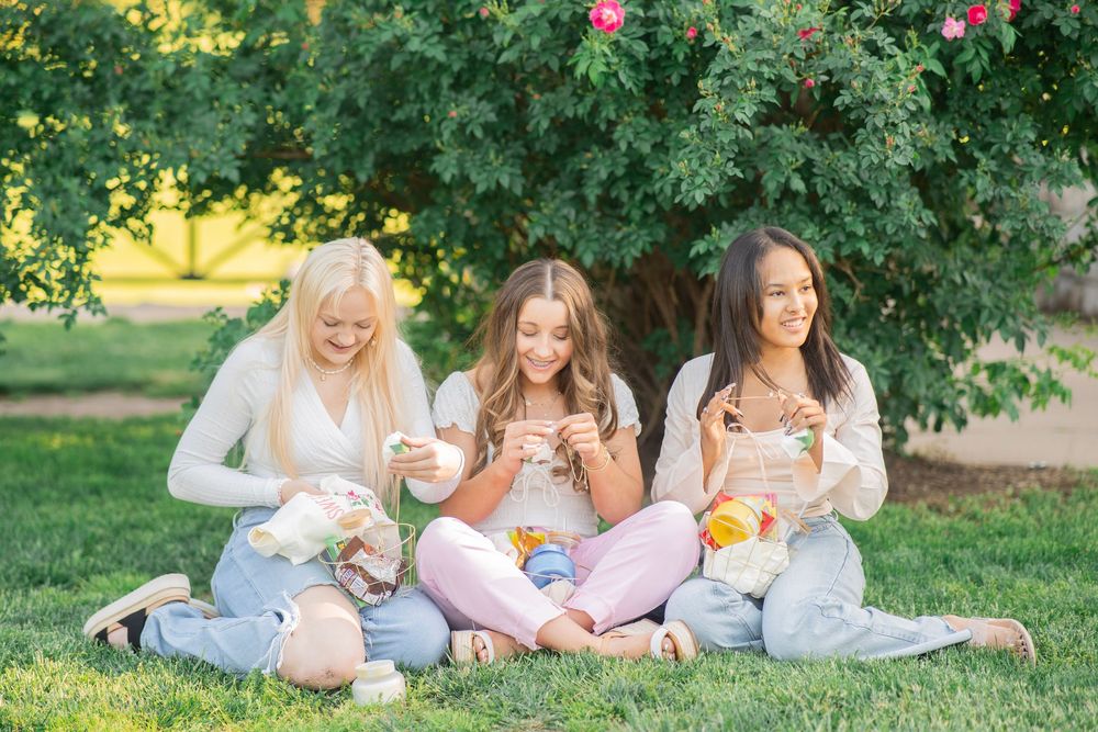 Three smiling teenage girls sitting on grass enjoying snacks and treats under a flowering tree.