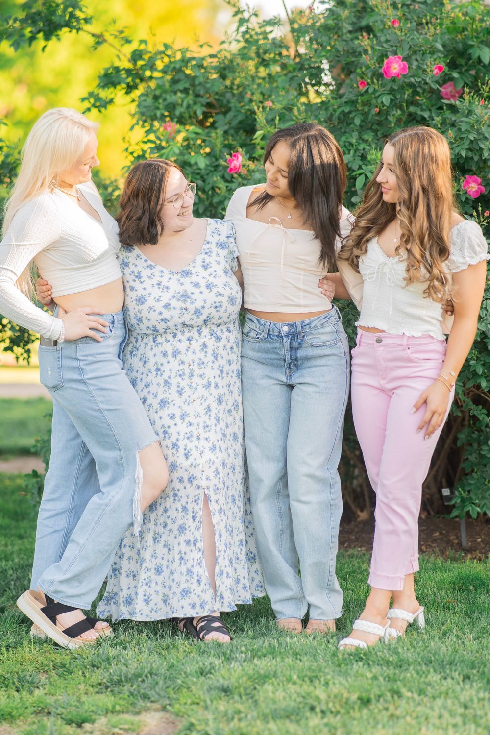 Four young women in casual spring outfits pose together laughing in a garden with pink roses.