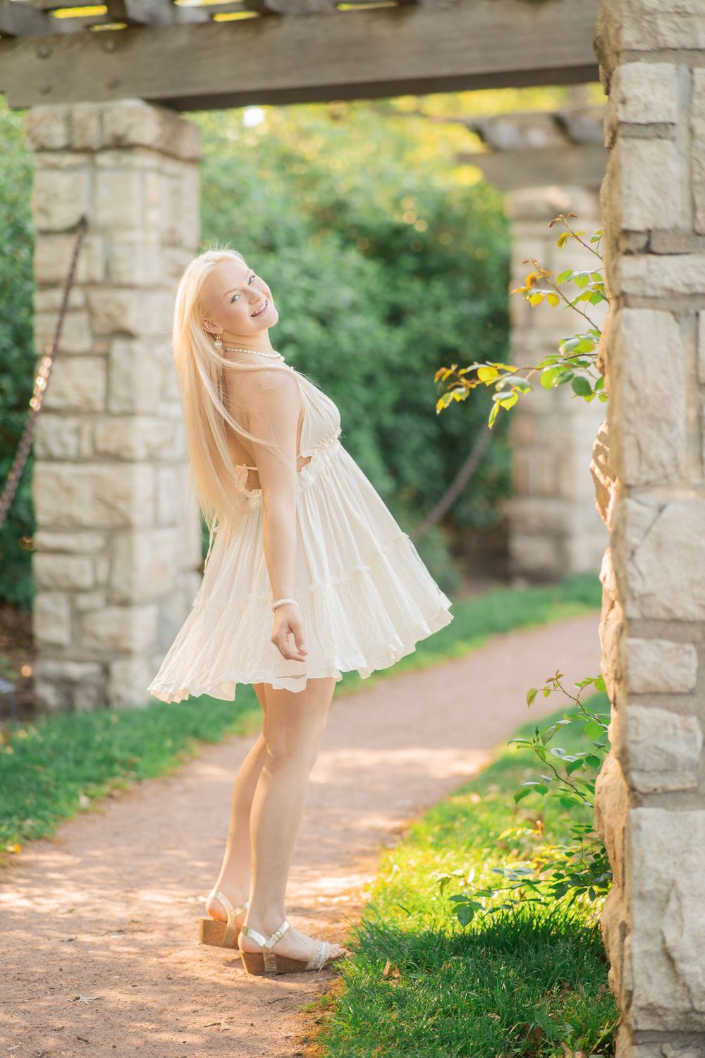 Young blonde woman in a white flowy mini dress smiling under a stone pergola in a garden setting.