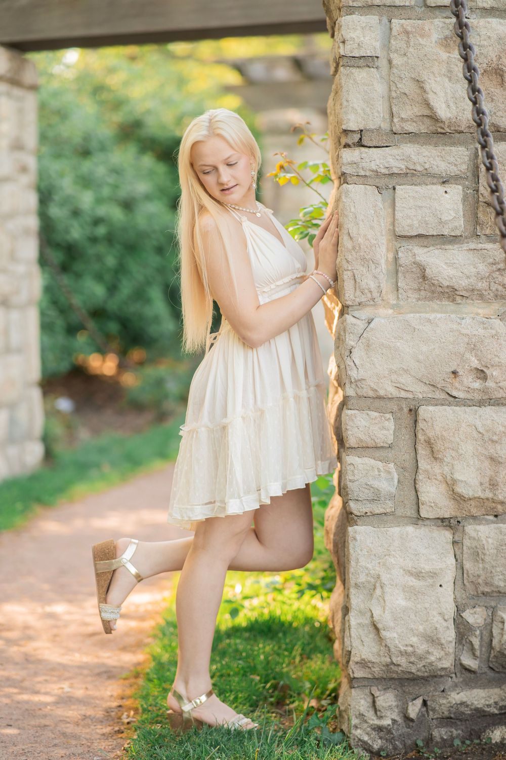 Blonde teenage girl in a cream ruffled dress posing beside a stone pillar in a sunlit garden.