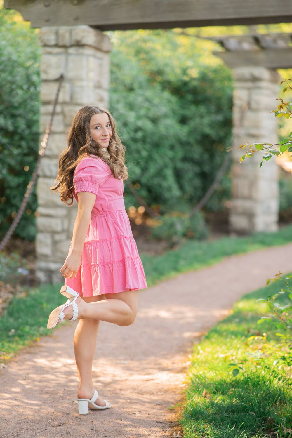 Teen girl in pink dress holding shoes, posing on garden path near stone pergola.