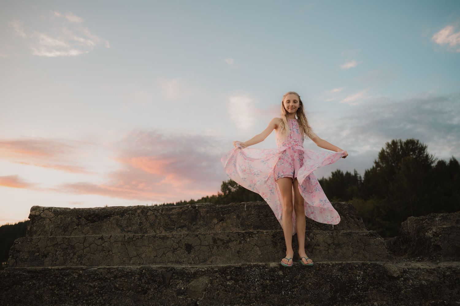 A person in a flowing pink dress stands gracefully on stone steps against a sunset sky with clouds.