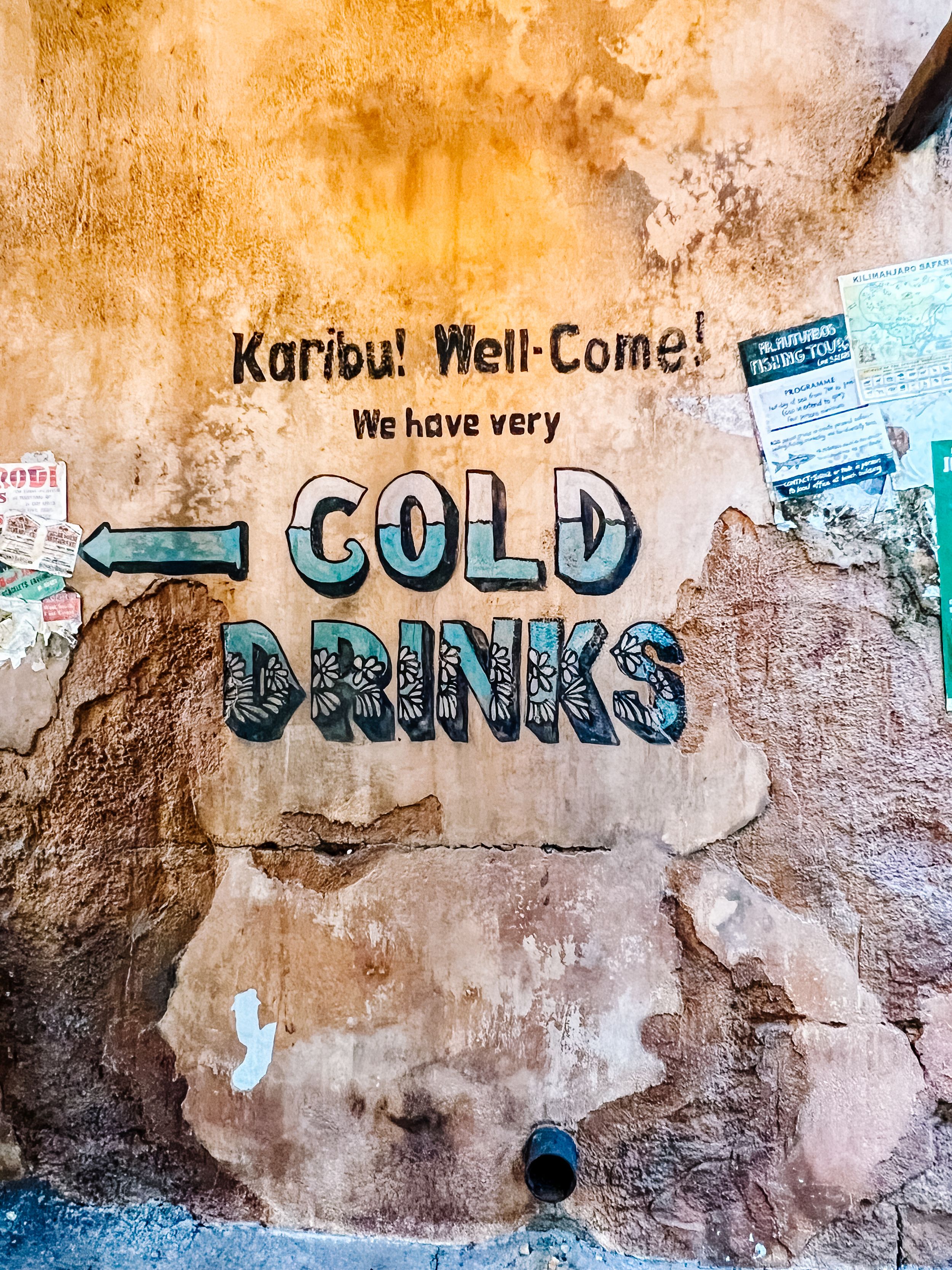 Hand-painted rustic wall sign displaying 'Karibu! Well-Come!' and 'COLD DRINKS' text in blue letters on weathered surface.