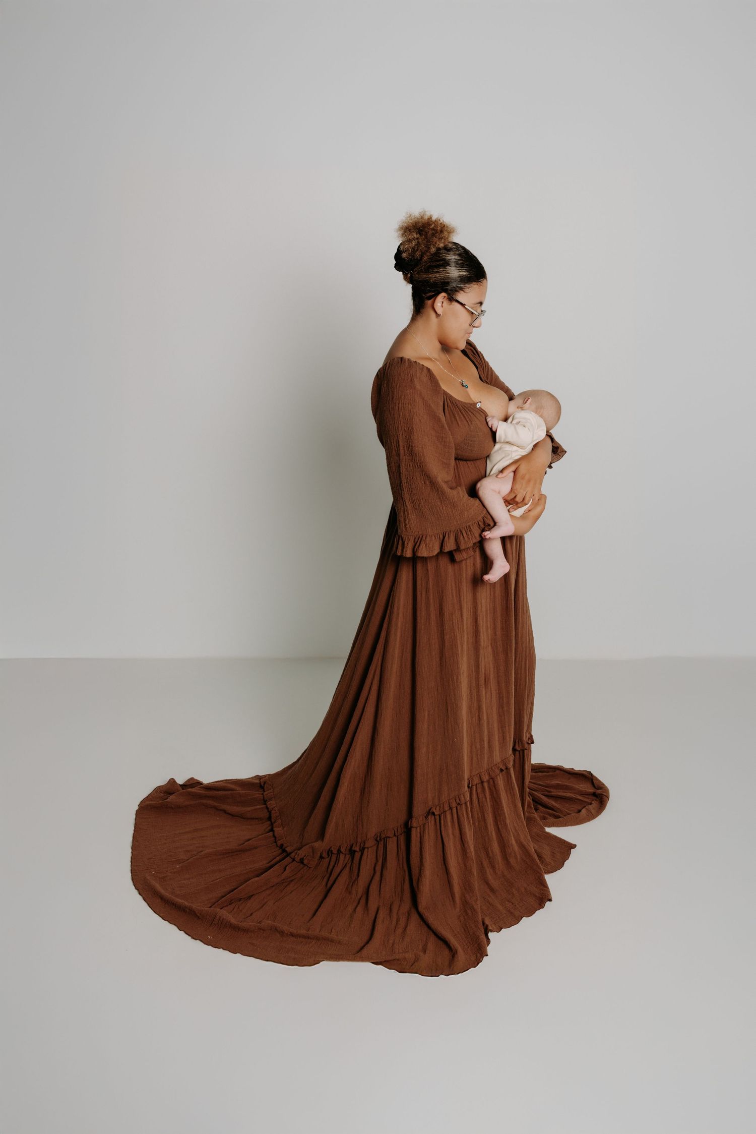 A pregnant woman in a flowing white lace dress stands on a sandy coastal path with ocean cliffs in the background.