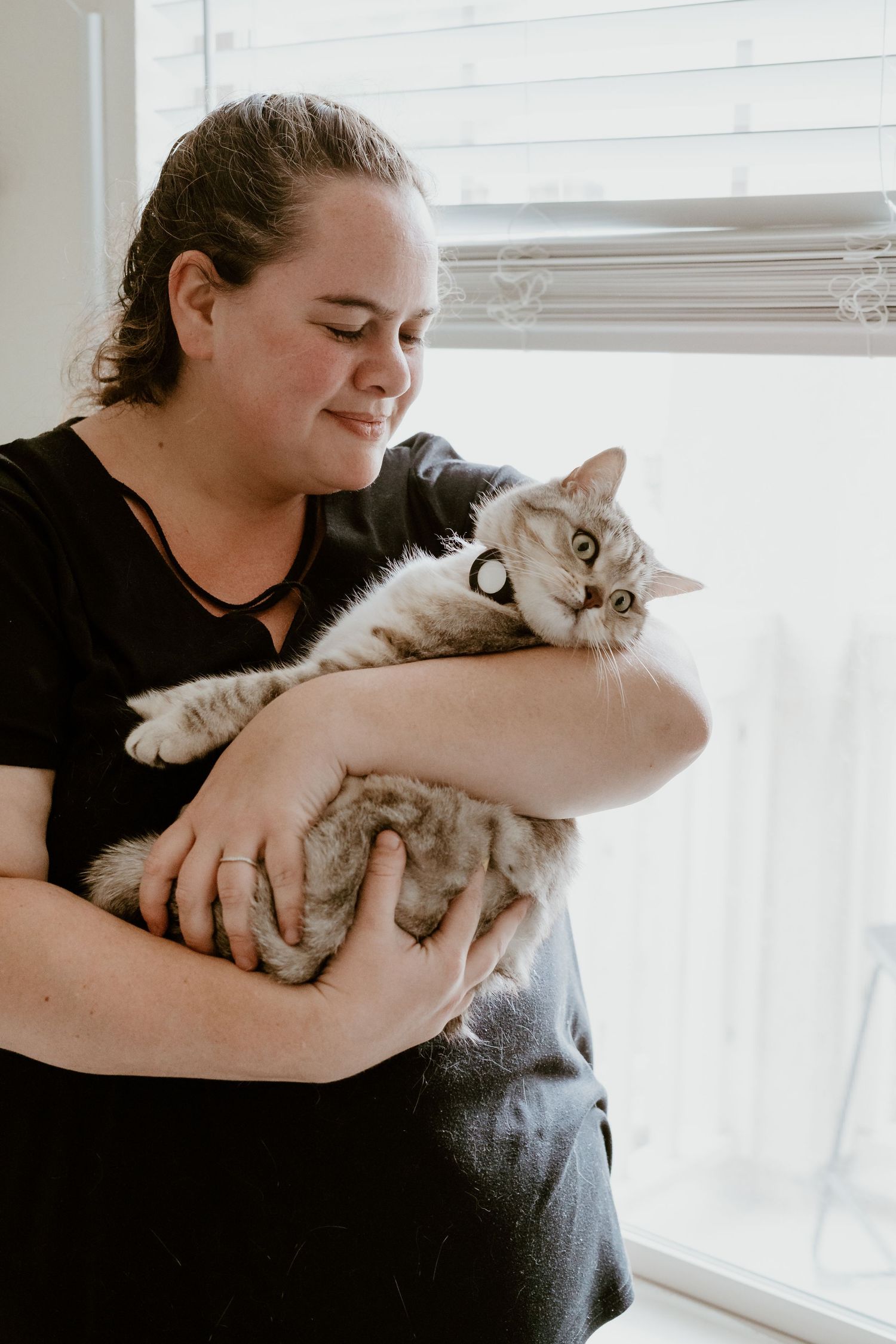 A loving moment between an owner in a black shirt holding a relaxed grey and white cat by a bright window.