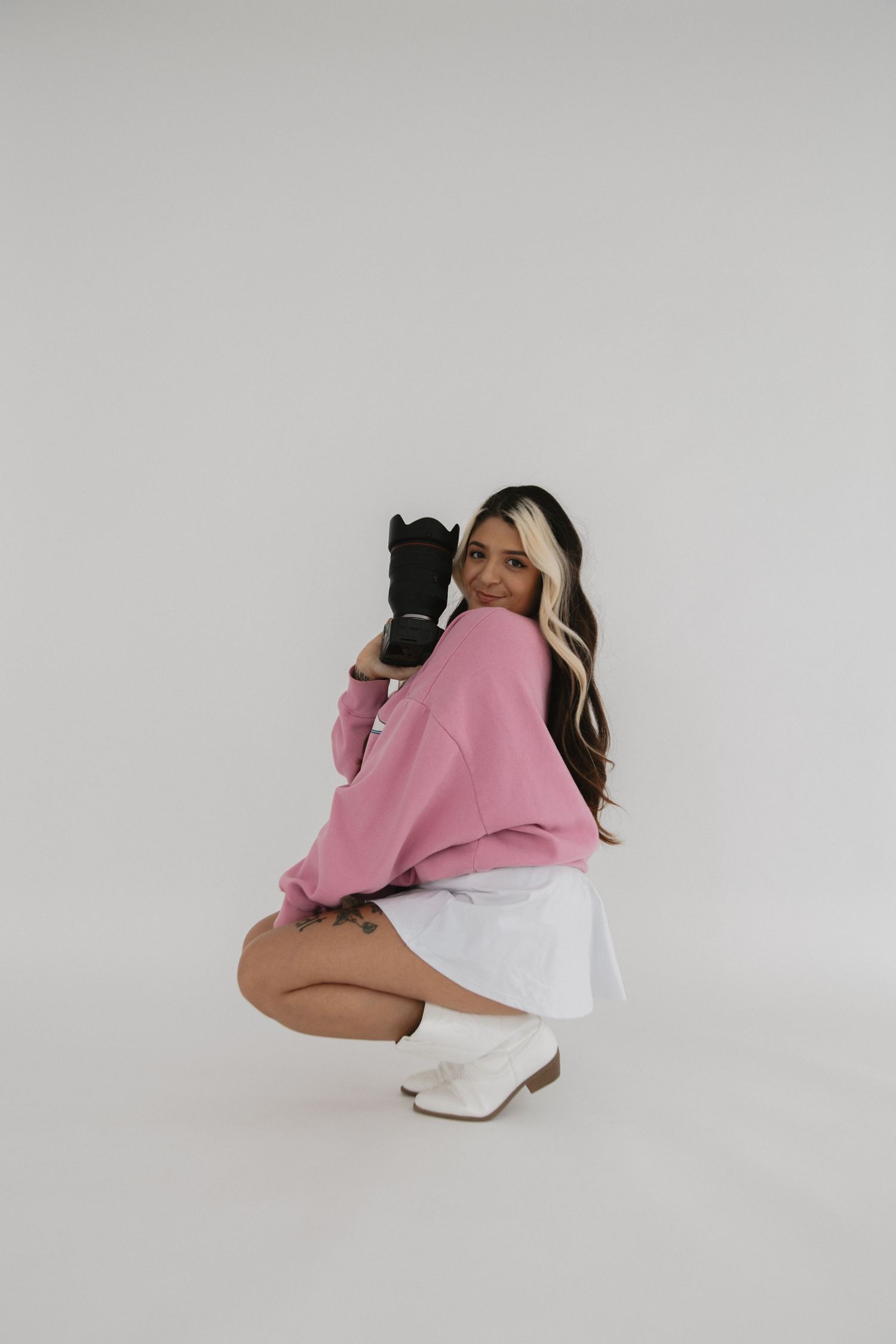 Photographer in a rustic sweater playfully posing with camera against white wall with air plants.