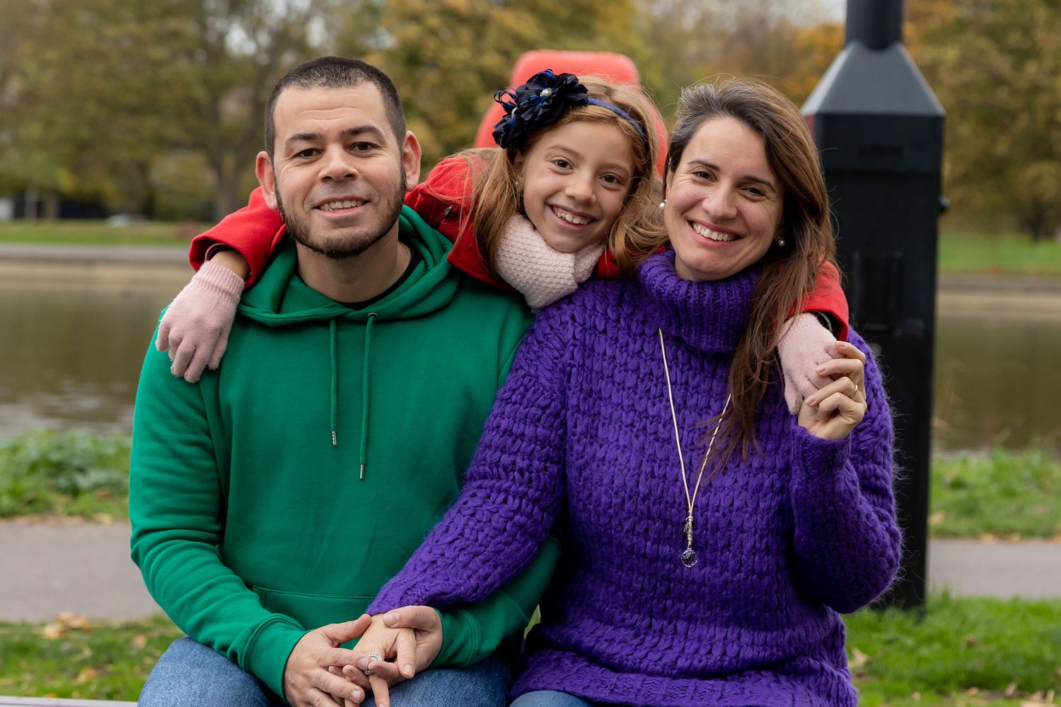 Three friends sitting together outdoors wearing colorful sweaters on an autumn day by a lake.