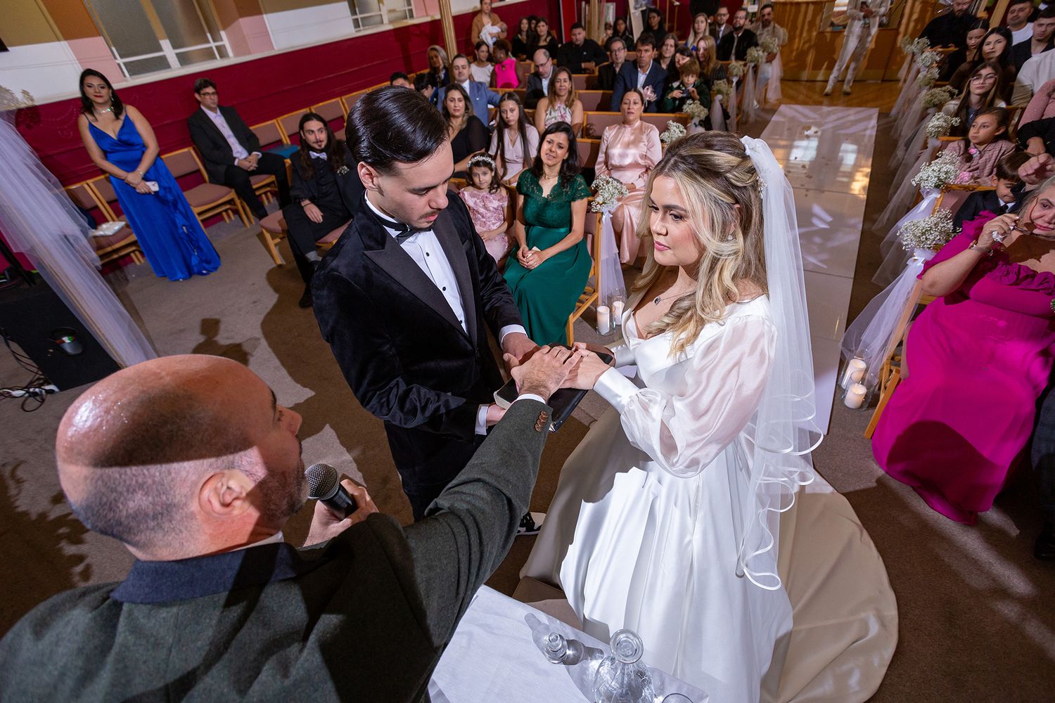 Wedding ceremony captured from high angle showing couple exchanging rings surrounded by guests in indoor venue.
