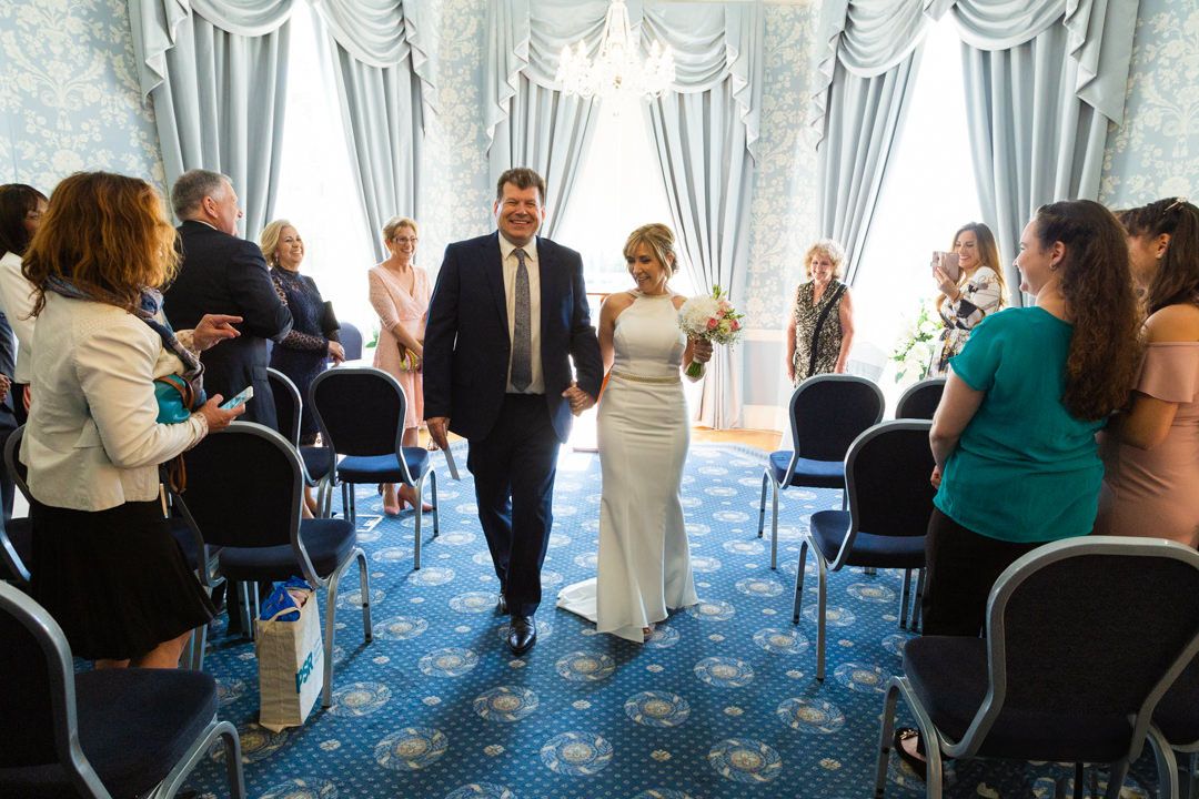 A couple walks down the aisle in a blue-carpeted room with white drapes as wedding guests look on and take photos.