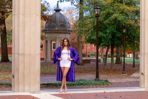 A person in a purple dress poses between stone columns on a sunny autumn day at a university campus.