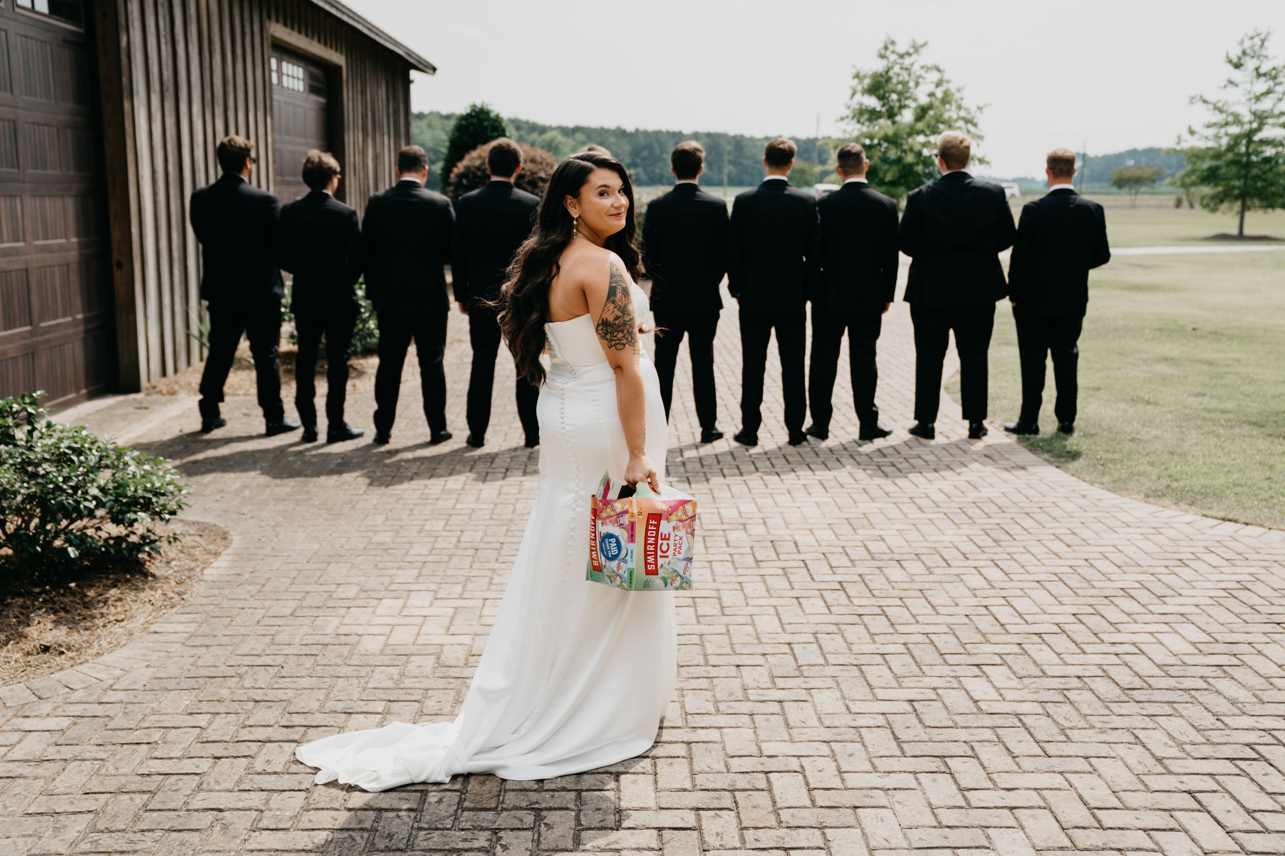 Bride in white gown holds colorful clutch while groomsmen in black suits stand facing a barn at an outdoor wedding.