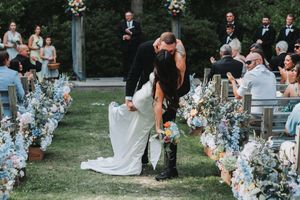 Bride and groom share a romantic dip kiss at outdoor wedding ceremony lined with blue and white floral arrangements.