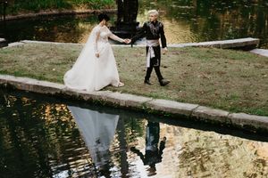 A couple in wedding attire walks along a pond's edge with their reflection visible in the calm water.