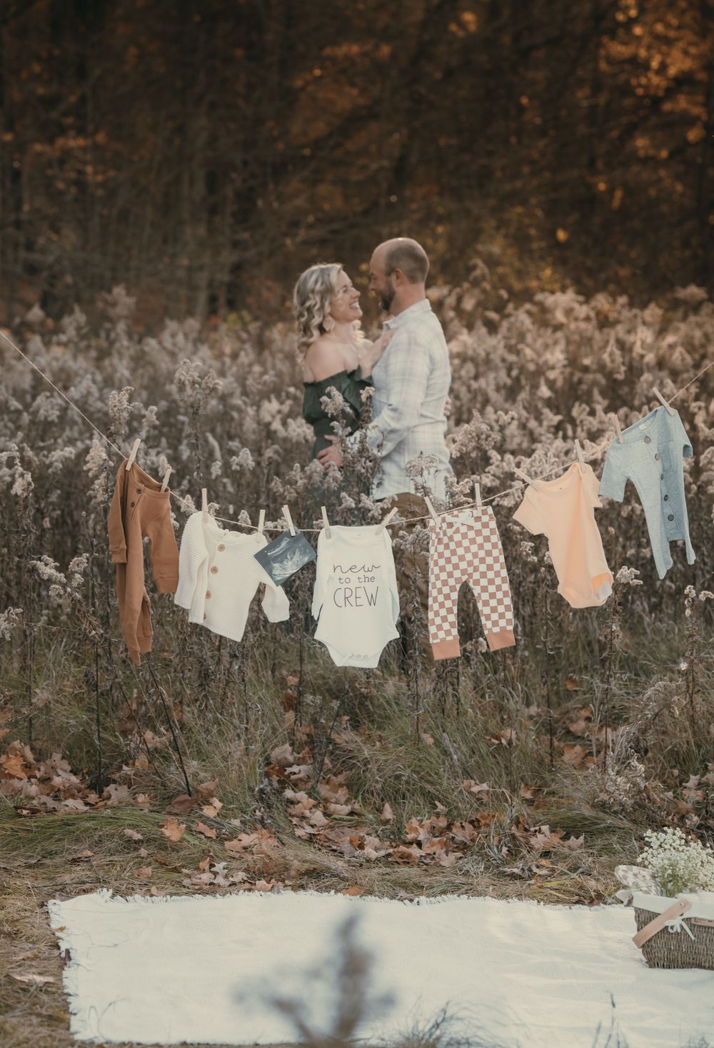 A couple embraces in front of a clothesline displaying baby clothes in an autumn forest setting.